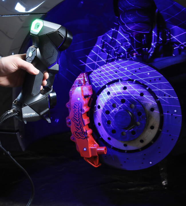 Technician using a Creaform HandySCAN 3D scanner to capture detailed 3D measurements of a car brake system, with projected laser grids visible on the components.