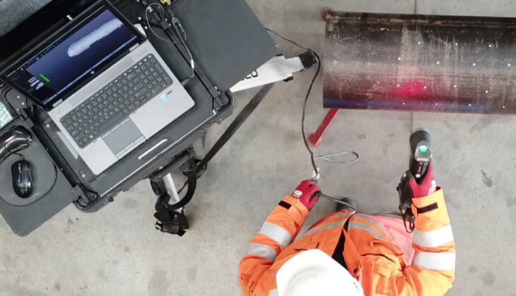 Overhead view of a technician in high-visibility gear using a Creaform 3D scanner to inspect a cylindrical industrial pipe, with a laptop displaying scanning data in real time.