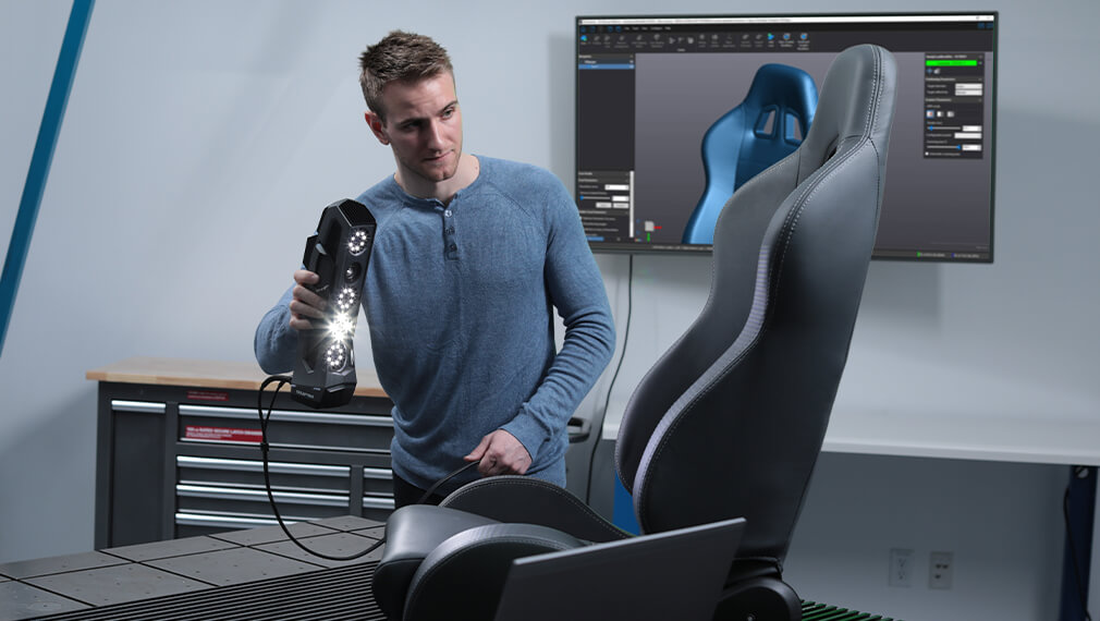Engineer using a Creaform HandySCAN 3D scanner to capture the geometry of an ergonomic car seat, with a 3D model displayed on a screen in the background.
