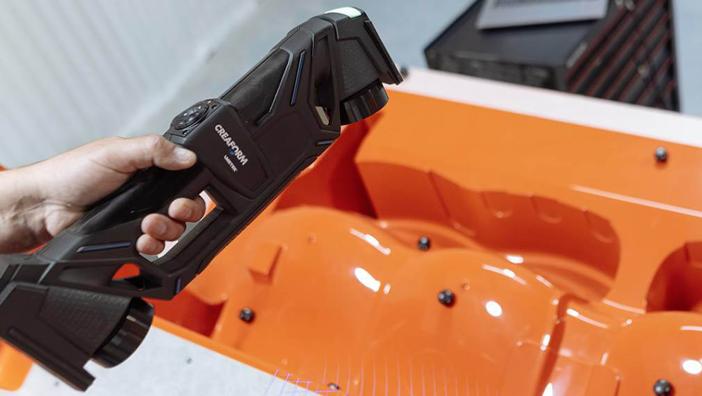 Hand holding a Creaform HandyPROBE portable probing device, inspecting an orange industrial mold for precise measurements in a quality control process.