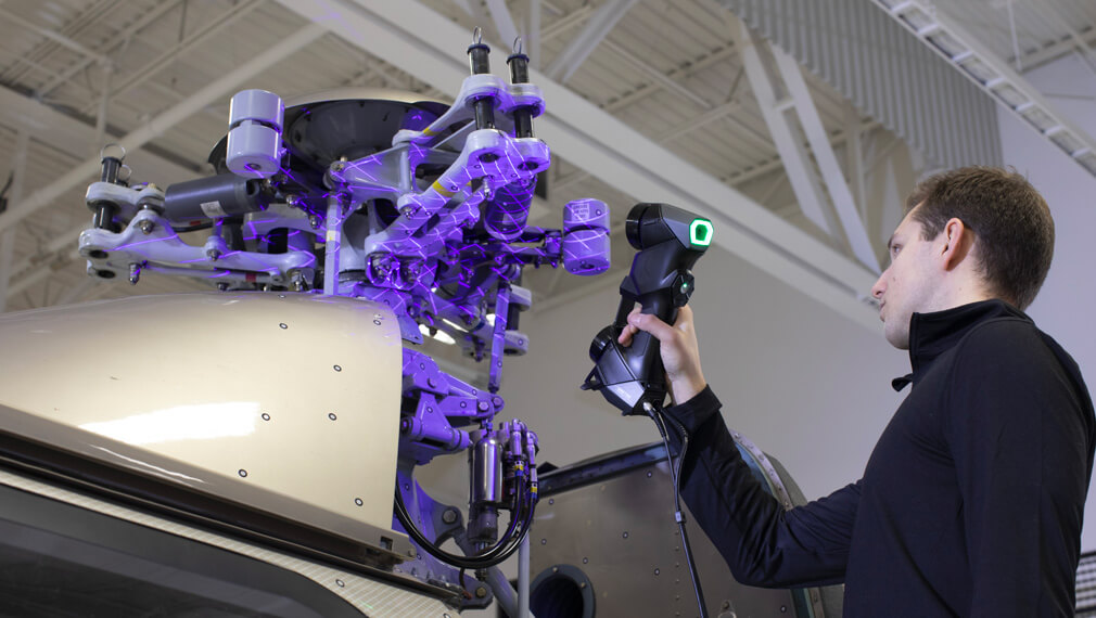 Technician using a Creaform HandySCAN 3D scanner to inspect the rotor assembly of a helicopter, with projected laser grids highlighting the scanned surfaces.